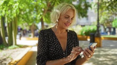 Ağaçların ve binaların bulanık arka planında gölgeli şehir parkında gülümseyen bir kadın, siyah-beyaz benekli elbise, kısa sarı saç ve inci küpeler takıyor..