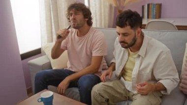 İki adam, muhtemelen kardeşler, mikrofon olarak bir TV kumandası kullanarak şarkı söylüyorlar. Parlak ve derli toplu bir oturma odasında, arka planda bir bardak ve kitaplarla birlikte rahat bir koltukta oturuyorlar..