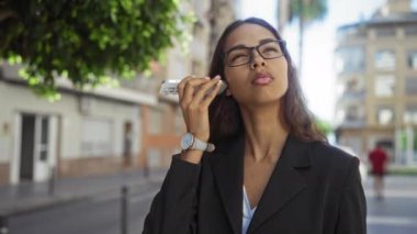 Şehir caddesinde cep telefonuyla konuşan, gözlüklü, siyah takım elbiseli, şehir binaları ve ağaç yapraklarıyla çevrili, düşünceli bir kadın.