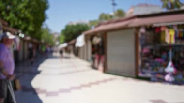 Kadın ve erkeğin yürüdüğü, konuştuğu, yaz güneşinin ve kırsal ortamın sergilendiği arka planda market tezgahları ve yeşilliklerin olduğu bulanık pazar manzarası.