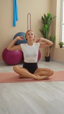 Spor salonunda yoga minderiyle esneyen, spor yapan, spor topları ve bitkilerle çevrili, sarı saçlı, siyah şortlu ve beyaz bluzlu bir kadın.