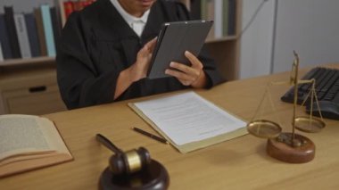 Belgeler, tokmak ve adalet terazileri masanın üstündeyken, mahkeme salonunda tablet ve klavyeyle çalışan siyah adli bornoz giyen bir adam.