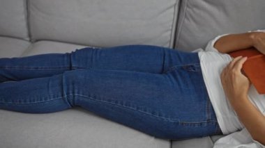 Oturma odasında göğsünde kitapla kanepede uyuyan olgun bir kadın. Evde rahat rahat giyiniyor.