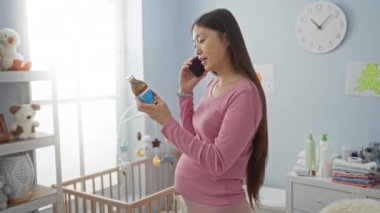 Pembe elbiseli hamile bir kadın, bebeğin huzurlu yatak odasında oyuncaklarla, beşiğiyle ve duvardaki saatle telefonda konuşurken şişeyi inceliyor..