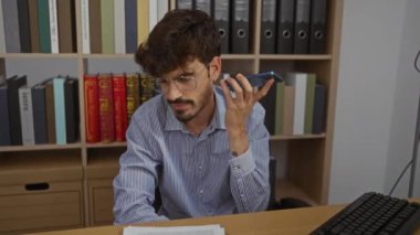 Ofiste akıllı telefon kullanan, gözlüklü ve çizgili gömlek giyen raflarda kitap ve klasörlerle çevrili genç bir adam bilgisayarla masa başında çalışıyor..