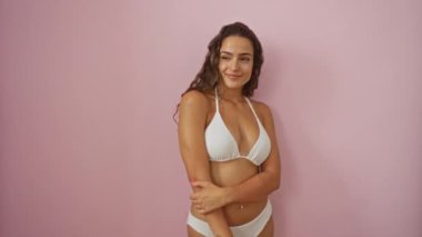 Bikini giyinmiş genç İspanyol kadın yan tarafa bakarken özgüvenle gülümsüyor, izole pembe arka planda gülüyor.