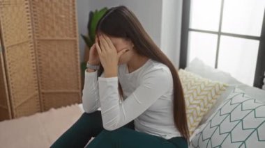 Yatak odasındaki yastıklardan ve pencerelerden süzülen doğal ışıktan endişeli bakışlarla yatakta oturan kadın..