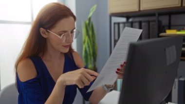 Kızıl saçlı kadın ofiste belge okuyor, gözlük takıyor ve gülümsüyor, açık mavi duvarlı ve arka planda yeşil bir çalışma ortamı sergiliyor.