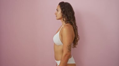 İspanyol bikinili genç bir kadın, özgüvenle poz veriyor, pembe arka planda doğal bir şekilde gülümsüyor.