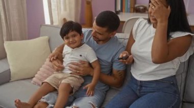 Aile, evdeki kanepede televizyon seyrediyor. Uzaktan kumandayı elinde tutan erkek ve kadınla kucaklaşıyor.