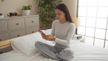 Endişeli bir kadın yatak odasında oturmuş elinde cep telefonuyla evde duygusal bir tepki verirken endişeli görünüyor..