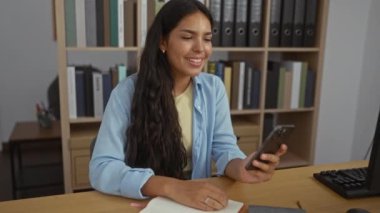 Ofiste telefonuna bakarken gülümseyen bir kadın. Etrafı kitaplarla ve kitaplarla çevrili. Bu da verimli bir çalışma ortamı olduğunu gösteriyor..