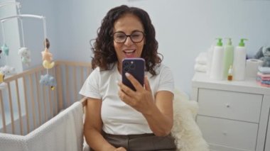 Sıcak yatak odasında bebek beşiği ve oyuncaklarıyla gülümseyen bir kadın, ev ihtiyaçlarıyla çevrili, sıcaklığını ve aile bağlarını içeride iletiyor..