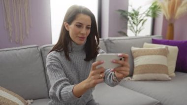Kadınlar rahat oturma odasındaki kanepede keyifli bir ifadeyle gri kazak giyerek mobil oyun oynamasını kutlarlar..