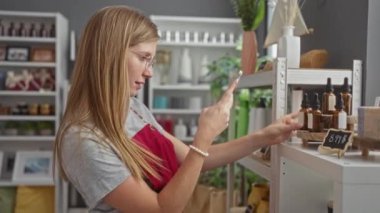 Young, attractive, blonde woman taking photos indoors with her phone in a beautifully decorated home decor store showcasing various items on shelves