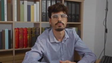 Ofiste oturan sakallı ve gözlüklü genç adam etrafı kitaplarla ve dosyalarla çevrili kameraya gülümsüyor.