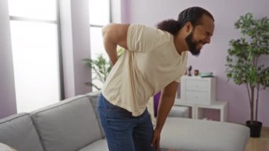 Oturma odasında sırt ağrısıyla boğuşan bir adam modern apartman dairesinde günlük giysiler, bitkiler ve kanepe giyiyor..
