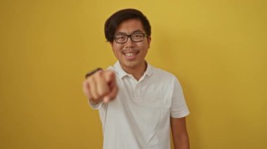 Genç adam kameraya neşeli bir ifadeyle bakıyor, parlak sarı bir arka planda gözlük takıyor.