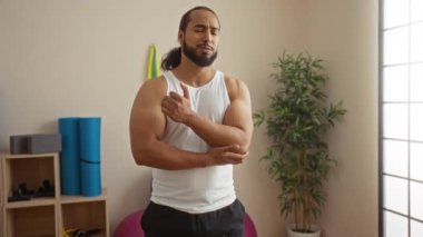 Spa odasında sakallı bir adam endişeli bir bakışla dirseğini tutuyor. Yoga minderleri ve saksı bitkileriyle çevrili. Sağlık durumundaki rahatsızlık veya acıyı gösteriyor..