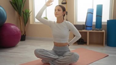 İspanyol kadın, spor salonundaki yoga minderinin üzerinde geriniyor ve arka planda egzersiz ekipmanları var.