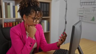 Ofiste çalışan bir kadın bilgisayar başında otururken akıllı telefon kullanıyor pembe ceket ve gözlük takıyor, etrafı kitaplarla ve çizelgelerle çevrili.