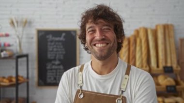 Önlüklü gülümseyen genç adam, ekmek somunlarıyla dolu bir fırında, karatahta menüsünün önünde poz veriyor.