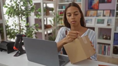 Genç bir kadın ev dekorasyonu mağazasında bir paket hazırlıyor. Masasında dizüstü bilgisayar ve barkod tarayıcısı var.