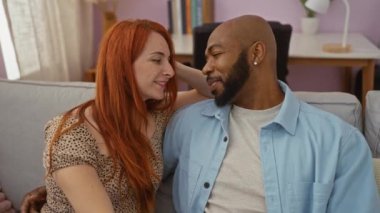 Irklar arası bir çift evdeki bir kanepede birbirlerine gülümseyip kucaklaşıyorlar. Sıcak bir oturma odasında sevgi ve mutluluk gösteriyorlar.