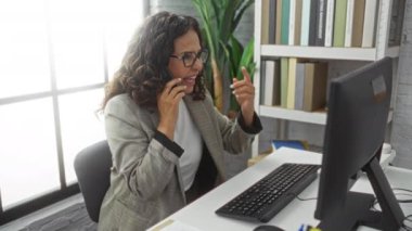 Modern ofiste telefonla konuşan bir kadın bilgisayarla uğraşırken, kitaplar ve bitkilerle çevrili, iç mekanı geliştiren doğal ışık ile profesyonel bir atmosfer sergiliyor..