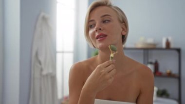 Güzeller güzeli bir kadın spa odasında kısa sarı saçlı ve sağlık merkezinde havlu giymiş bir yüz silindiri kullanıyor.