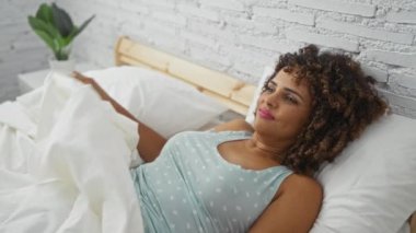 Genç bir kadın yatak odasında kıvırcık saçlarıyla uyanıyor ve sabah gülümsüyor, açık mavi bir bluz giyiyor, beyaz bir yatak örtüsünün üzerinde beyaz tuğladan duvarları olan bir odada yatıyor.