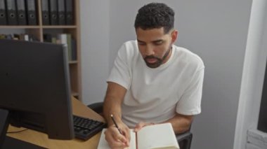 Bir ofiste çalışan genç bir adam, bir dizüstü bilgisayarla klasörlerin önünde otururken bir not defterine yazı yazıyor.