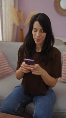 İspanyol kadın gülümsüyor ve rahat bir oturma odasında telefon kullanıyor modern iç dekorasyonu ve rahat koltuğu var.