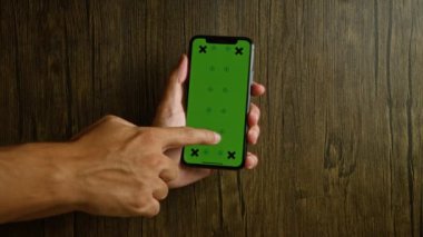 Adam ahşap bir arka planda otururken yeşil ekranlı akıllı telefondan el ve parmaklarına odaklanıyor.