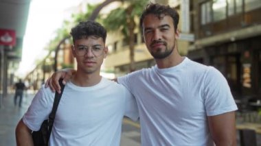 İki İspanyol adam, muhtemelen bir baba ve oğul, beyaz gömlekler giyiyorlar, gülümsüyorlar ve arka planda binalar olan bir şehir sokağında kucaklaşıyorlar..