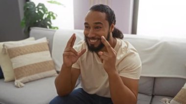 Oturma odasındaki başarıyı kutlayan adam parmaklarını çapraz yapıp yumruklarını kaldırmış, evinde neşe ve heyecan gösterisi yapıyor, rahat bir dekor ve doğal ışık sunuyor..