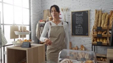 Bir pastanede oturan, önlük giyen ve kameraya güvenle gülümseyen bir kadın. Etrafı ekmek, hamur işleri ve pişmiş yiyeceklerle çevrili.