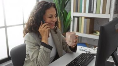 Elinde kahve fincanı tutarken telefonla konuşan olgun bir kadın, modern ofiste kitap rafları ve bilgisayarla masa başında oturuyor, profesyonellik ve samimiyet yayıyor..