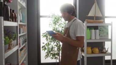 Önlüklü genç adam arka planda çeşitli eşya ve bitkilerle dolu bir mağazada tablet kullanıyor.