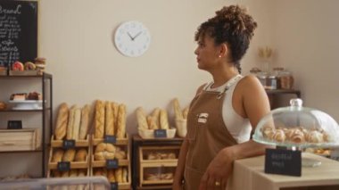Genç bir kadın pastanede çalışıyor. Arkasında ekmek ve hamur işleri var.