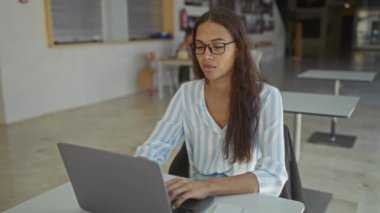 Dizüstü bilgisayarda çalışan bir kadın omuz ağrısı çekerken genç bir esmer modern bir iş ortamında duruşunu düzeltiyor.