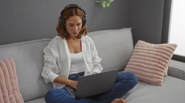 Kulaklıklı genç bir kadın evdeki bir oturma odasında kanepede otururken dizüstü bilgisayar kullanıyor.
