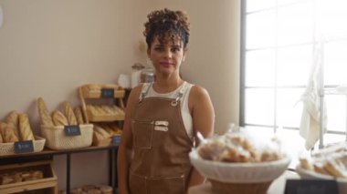 Fırında kolları çapraz duran bir kadın, önlük takıyor, arka planda ekmek rafları ve fırınlanmış mallarla dolu, pencereden gelen doğal ışıkla aydınlatılıyor.