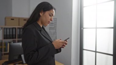Ofiste siyah ceket giyen, akıllı telefon kullanan, pencerenin yanında duran kitaplıkları ve arka planda bir masası olan İspanyol bir kadın.