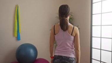 Spor salonundaki pembe tişörtlü kadın ciddi bir ifadeyle fitness toplarının yanında duruyor ve içeride direnç bantları takıyor..