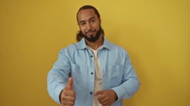 Sakallı genç bir adam, sarı duvarda izole edilmiş, mavi gömlekli, kendinden emin bir ifadeyle onaylamadığını gösteren baş parmak işareti yapıyor.