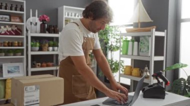 Ev dekorasyonu mağazasında çalışan genç bir adam, çeşitli iç dizayn ürünleriyle çevrili, dizüstü bilgisayar kullanıyor ve önlük giyiyor.