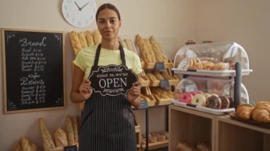 Fırında elinde ekmek ve pastalarla çevrili açık tabela tutan kadın, gülümsüyor ve dükkanın çeşitli fırınlanmış ürünlerle açık olduğunu gösteriyor.