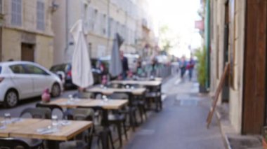 Marsilya 'da güneşli bir Fransız caddesinde masaları olan bulanık bir açık hava kafe sahnesi. İnsanlar geçerken, kentsel yaşamın canlı Avrupa atmosferini yakalıyorlar..