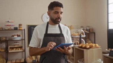 Pastanede arka planda raflar dolusu hamur işi olan tablet kullanan genç adam.